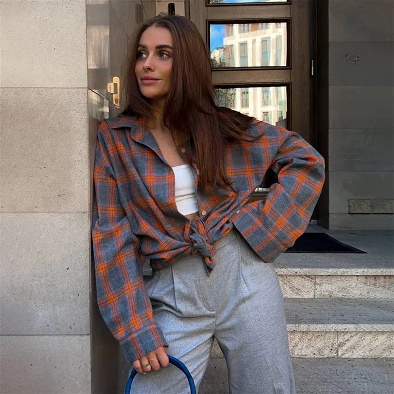 Woman wearing a plaid shirt and gray pants leaning against a building.