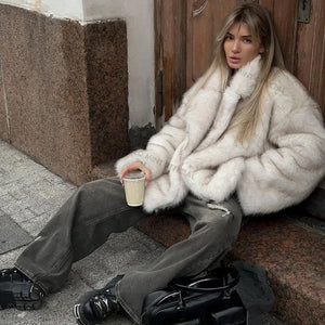 Person wearing a white fur coat sitting on a step holding a drink.