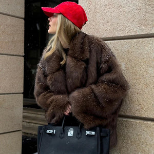Person wearing a brown fur coat and red cap, holding a black handbag against a stone wall.