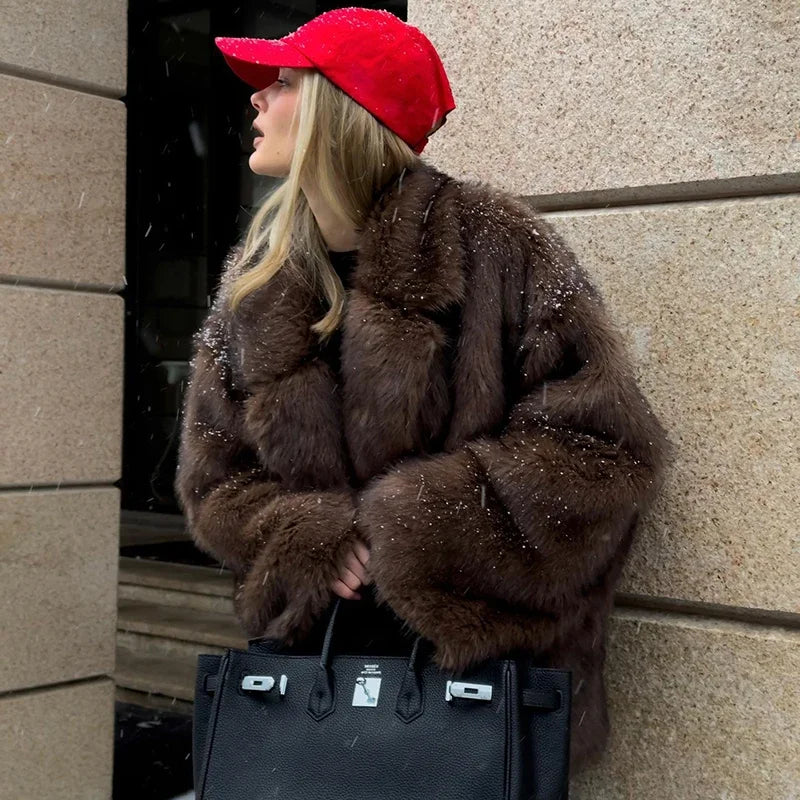 Person wearing a brown fur coat and red cap, holding a black handbag against a stone wall.