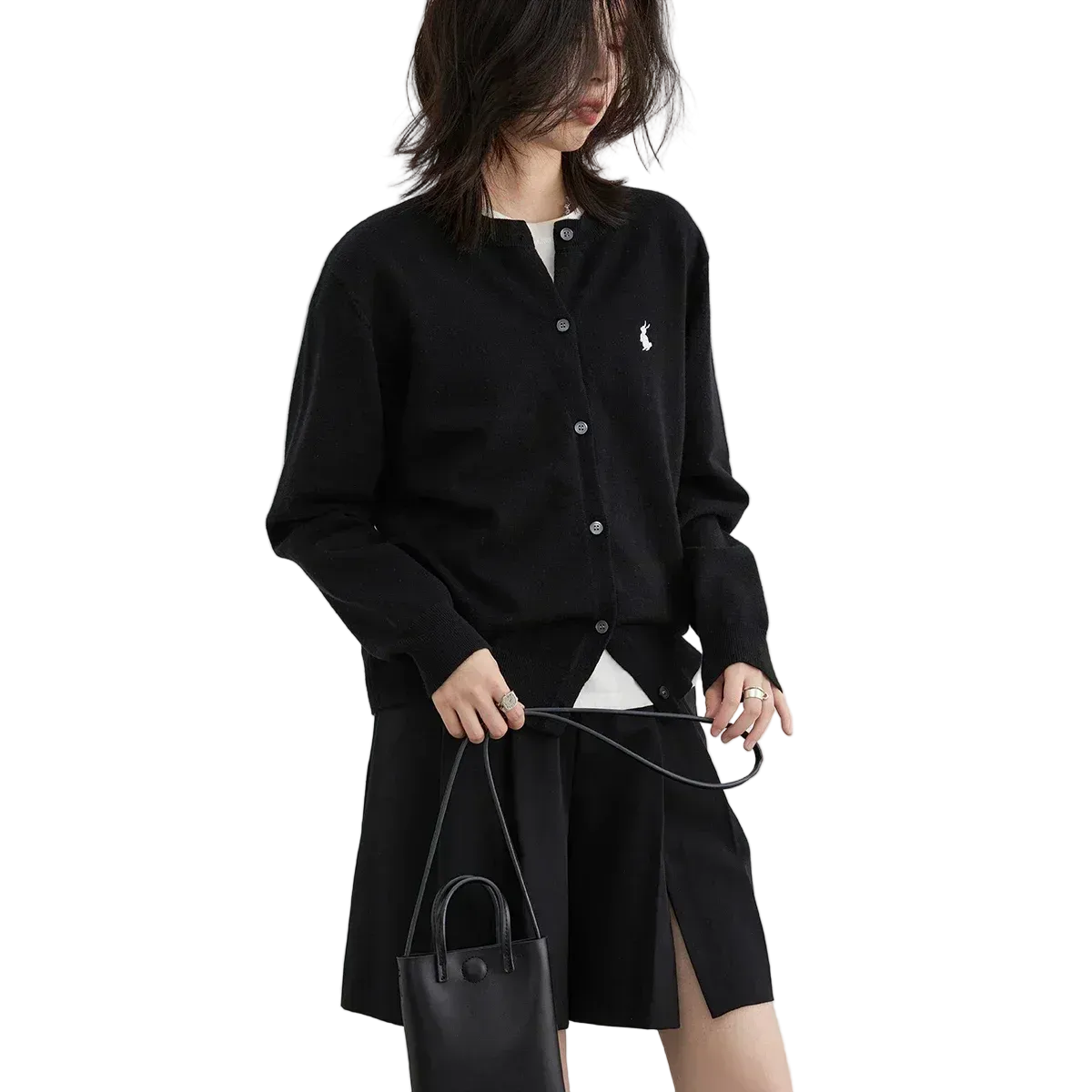 Person wearing a black cardigan with a brand logo, holding a black handbag on a white background