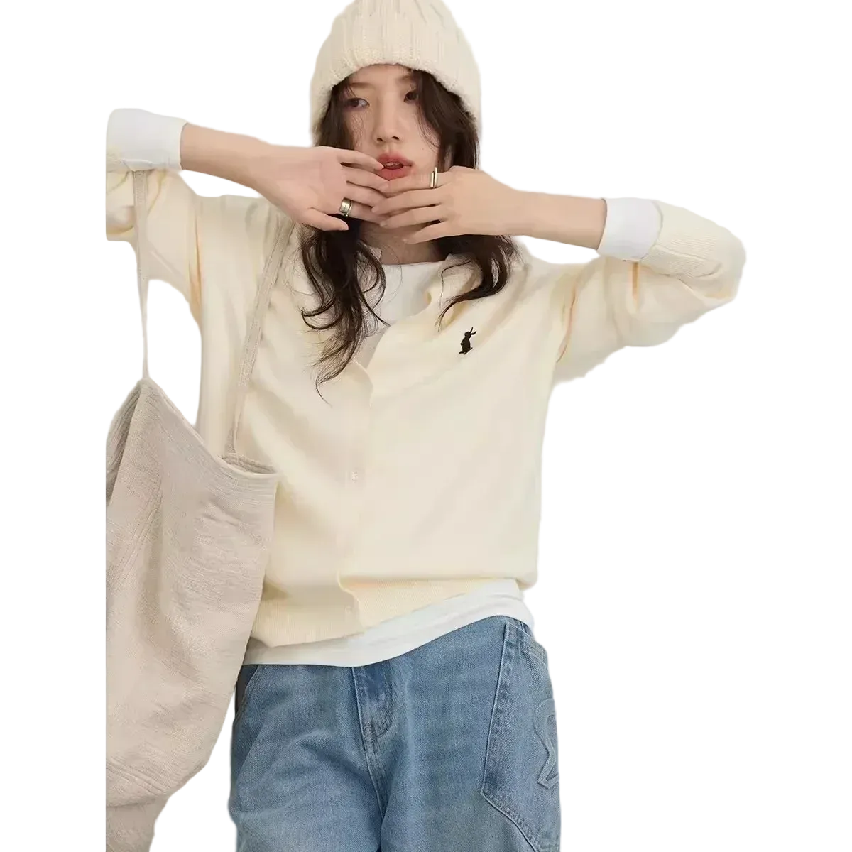 Person wearing a cream-colored sweater with a logo, light blue jeans, and a beige coat on a white background