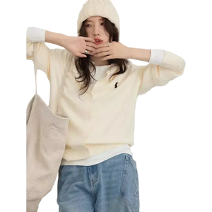 Person wearing a cream-colored sweater with a logo, light blue jeans, and a beige coat on a white background