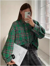 Person wearing a green plaid shirt holding a phone and paper in a room with a window.