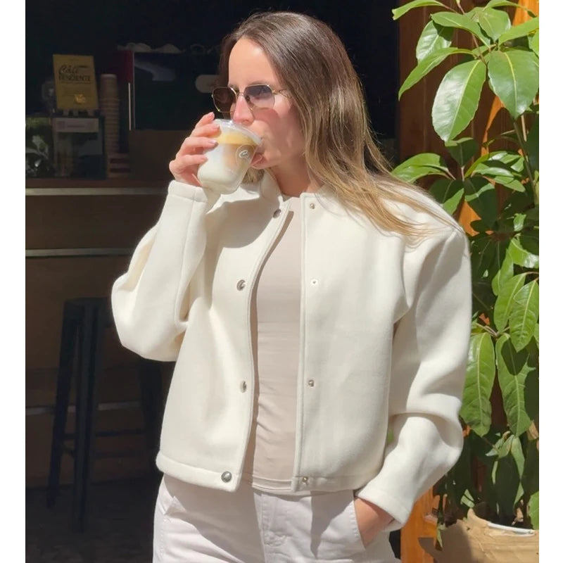 Woman in a white jacket drinking from a cup with a plant and indoor setting in the background