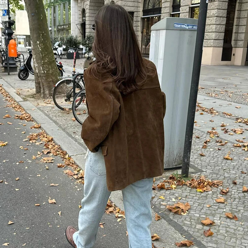 Person wearing a brown suede jacket and light blue jeans walking on a sidewalk with autumn leaves.