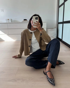 Person sitting on the floor taking a mirror selfie wearing a beige cardigan, white shirt, dark pants, and black studded flats.